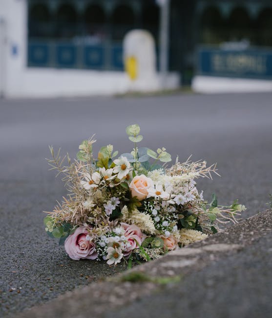 A delicate floral arrangement consisting of soft pink roses, white daisies, and light green foliage, including eucalyptus and dried grasses, lies on the pavement near a curb. The bouquet is loosely ga
