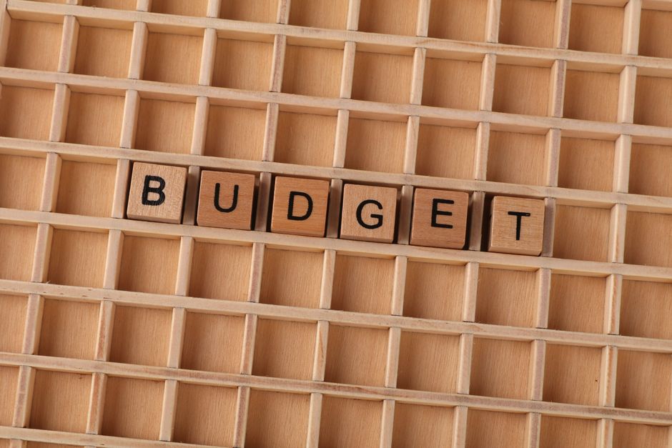 A close-up view of a light wooden gridboard with small square compartments, on which the word 'BUDGET' is spelled out using seven small wooden letter tiles with black uppercase letters. The background
