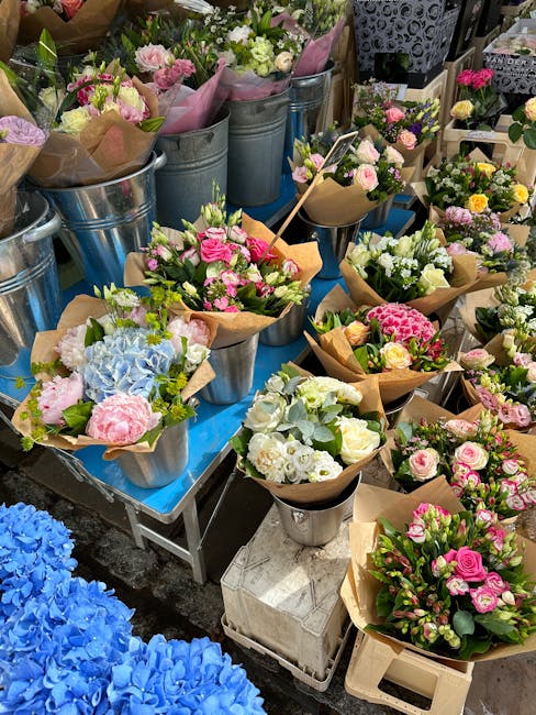 A vibrant display of freshly arranged flower bouquets and potted plants, featuring pink, white, and blue flowers including roses, hydrangeas, and lisianthus, set on a blue table at a florist stall. Th