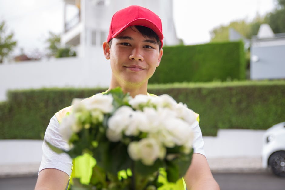 A smiling delivery person wearing a red cap and a neon yellow safety vest holds out a bouquet of white roses and green foliage towards the camera outdoors. The bouquet is fresh, with velvety petals an