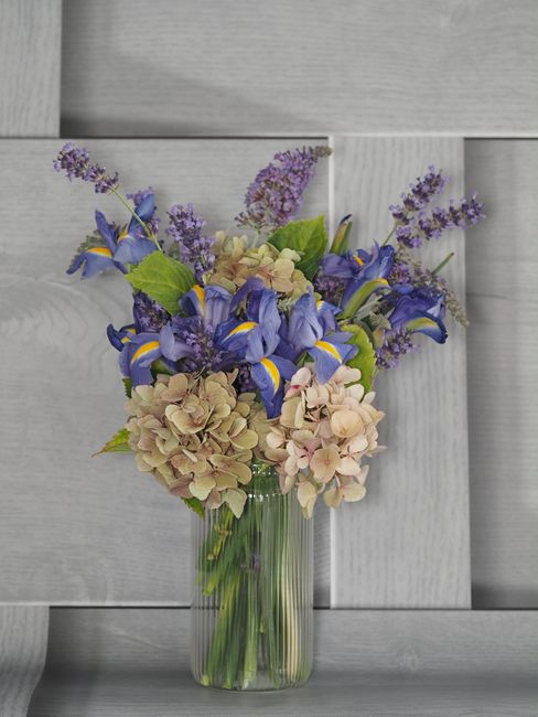 A vibrant floral arrangement in a clear glass vase featuring a mix of soft pink hydrangeas, purple lavender sprigs, and striking blue and yellow iris flowers. The bouquet is set against a light grey w