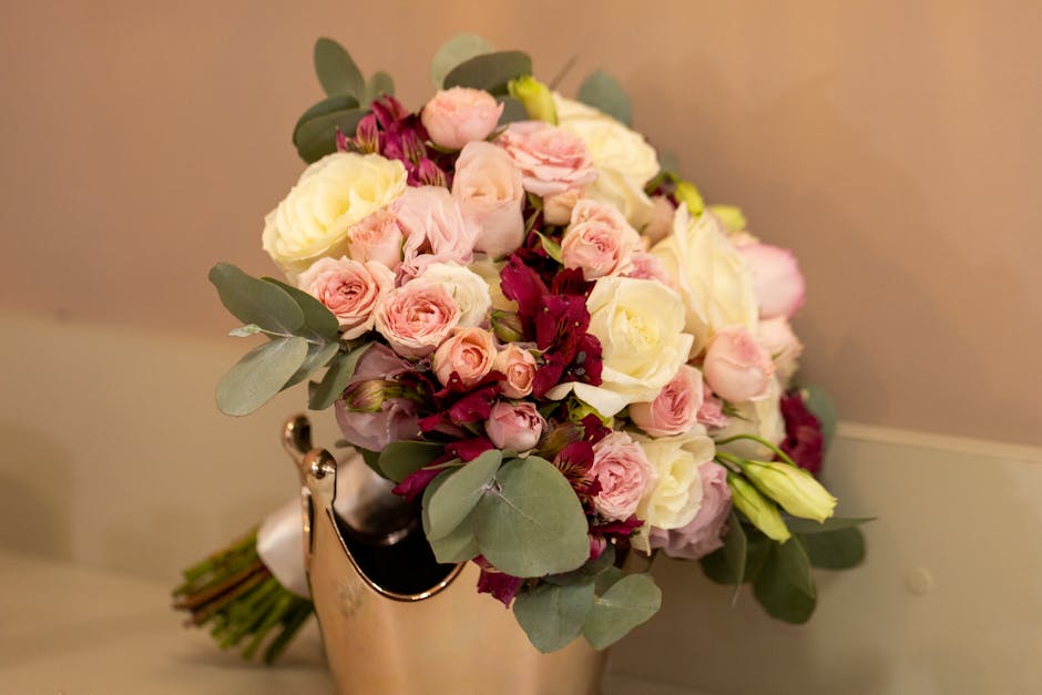 A floral bouquet arrangement featuring a mix of pale pink and cream roses, accented with small dark pink and burgundy blossoms, and lush green eucalyptus leaves. The flowers are densely clustered in a