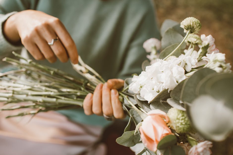 A florist from flowerdeliverykensalgreen.co.uk is assembling a floral gift, holding a bouquet composed of white stock flowers, soft pink roses, and delicate green chrysanthemums, surrounded by large, 