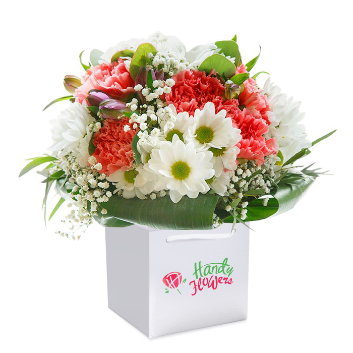 A beautifully-rounded bouquet rises elegantly from within a fresh white square gift bag, the whole ensemble front and centre upon a spotless white backdrop. This plush arrangement is artfully composed of coral-pink and vibrant red carnations-their plentiful ruffled petals adding volume and a striking visual softness. Sprinkled throughout are crisp white daisies with eye-catching lime-green centers, their playful faces creating a welcoming contrast. Wisps of delicate baby's breath thread their way delicately among the larger flowers, offering a gentle cloud-like finish that invites thoughts of peaceful moments in Kensal Rise. A careful scattering of unopened purple-tinged buds adds intrigue and a sense of unfolding beauty, emblematic of the ever-changing gardens around Ladbroke Grove. Generous, deep green foliage creates both support and a striking backdrop-the leaves broad, luscious, and immaculately arranged to frame the bouquet's lower edge. The entire piece is illuminated by soft, diffused light, ensuring that each hue and detail emerges with vibrancy and clarity. With its sturdy white ribbon handles, the gift bag is as practical as it is stylish, perfectly suited for delivery to local addresses or presentation at special gatherings. This image reflects not only the freshness and artistry found at Flower Delivery Kensal Green, but also the sense of connection offered by sending flowers for life's meaningful occasions-a tribute to new beginnings and cherished memories alike.